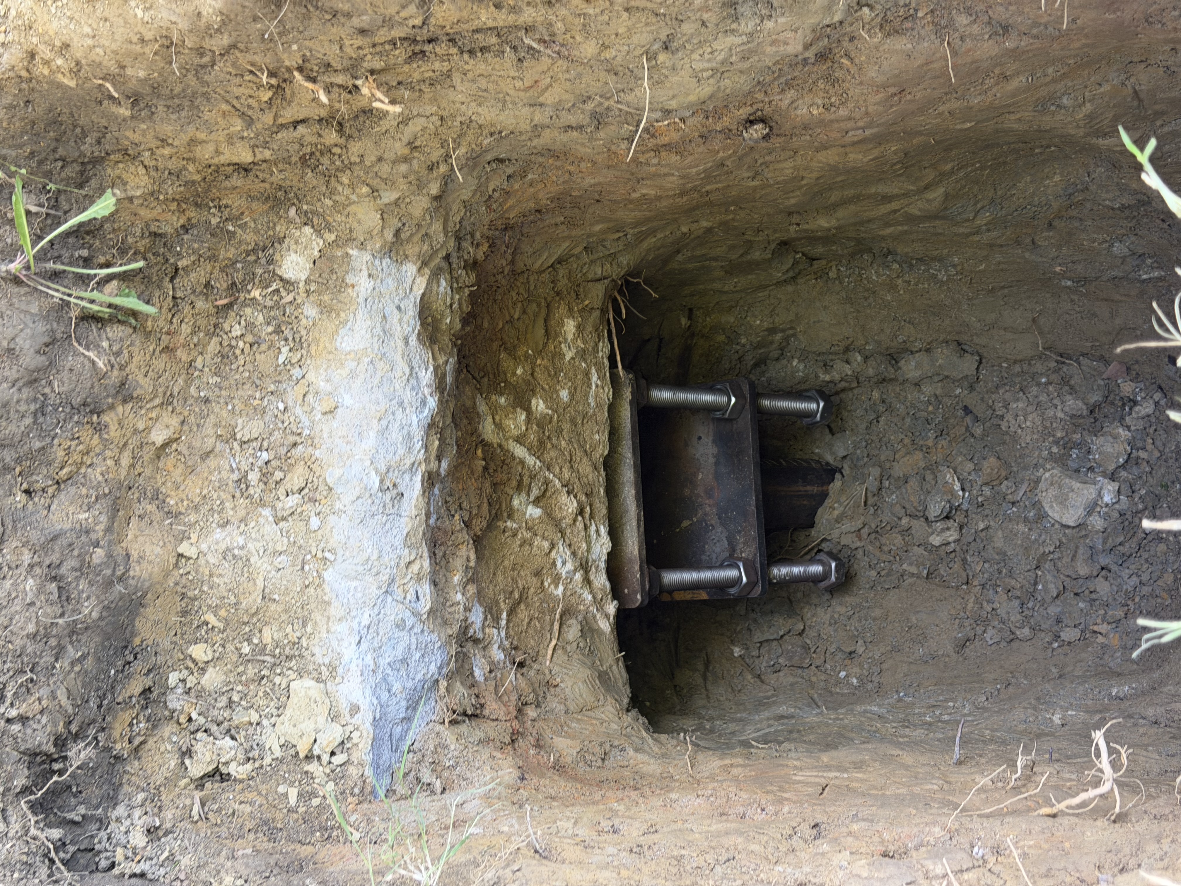 Steel pier foundation bracket bolted to a concrete footing inside an excavated hole, showing galvanized steel hardware secured against the foundation wall in North Texas clay soil